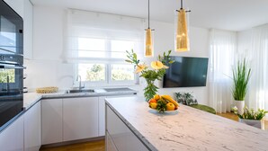 Eine moderne Küche mit weißen Schränken, einer Marmorarbeitsplatte, einer Spüle und einer Obstschale. Es gibt hängende Pendelleuchten und ein Flachbildfernseher an der Wand.