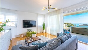 Ein modernes Wohnzimmer mit grauem Sofa, einem Flachbildfernseher, einer Essecke mit Tisch und Stühlen und großen Fenstern mit Meerblick.