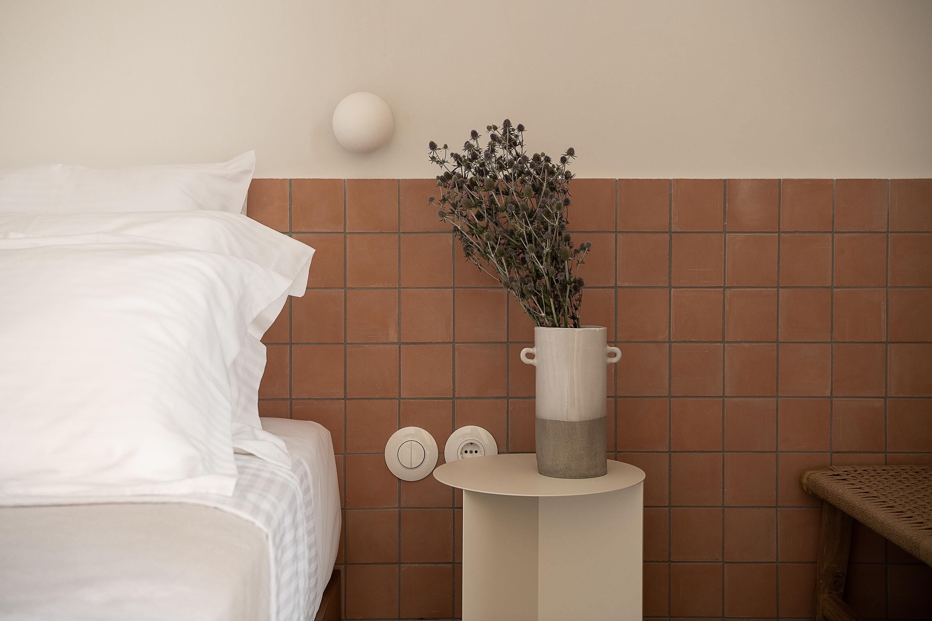 Una cama bien hecha con sábanas blancas, un florero con flores secas sobre una mesita y una pared con baldosas marrones.