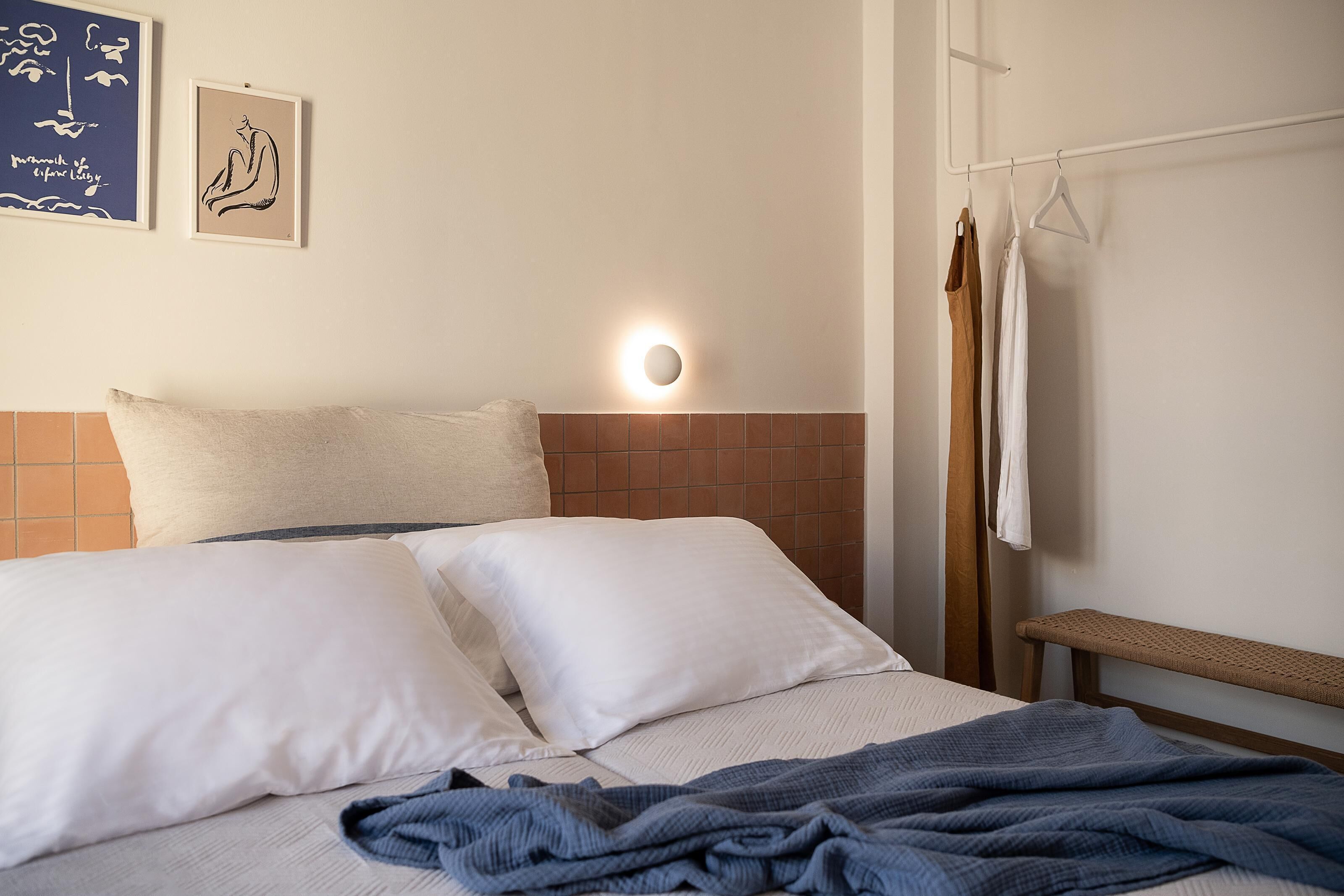 A neatly made bed with white and beige bedding, a wall-mounted light, and a bench.