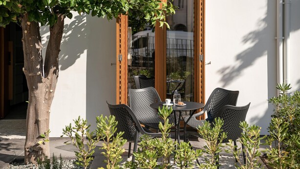 A patio with a table and chairs, a tree, and a building with a large window. - Lofos Casteli Sunlight (Chania)