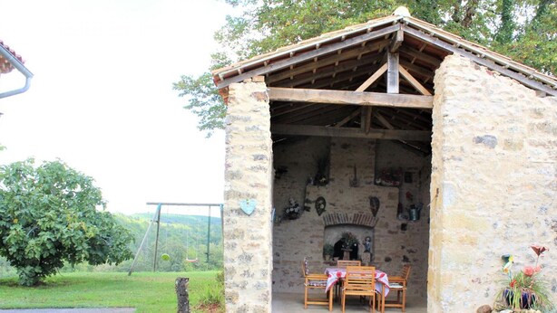 A stone building with a wooden roof, a fireplace, and a table with chairs outside.