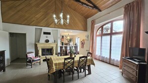 A spacious living area with a high ceiling, a fireplace, a dining table set for four, and a television on a dresser.