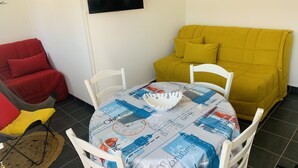 A small, cozy room with a yellow sofa, a red chair, a white table with a patterned tablecloth, and a flat-screen TV mounted on the wall. - Studio cabin with sea view and garage in Fort-Mahon plage (Fort-Mahon-Plage)