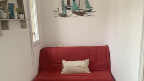 A red sofa with a cushion, a wall-mounted sailboat decoration, a woven rug, and a tiled floor. - Studio cabin with sea view and garage in Fort-Mahon plage (Fort-Mahon-Plage)