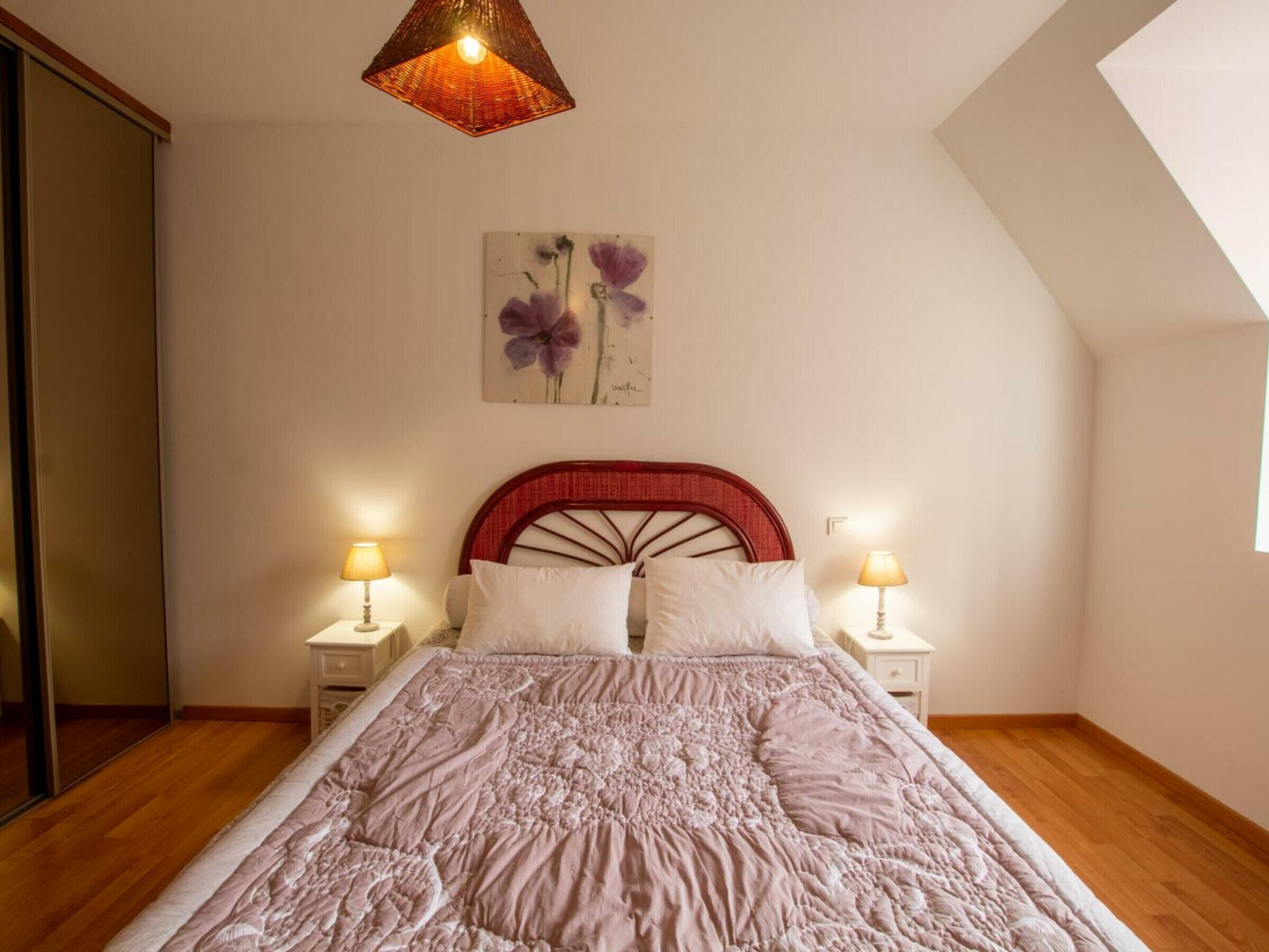 A bedroom with a wooden floor, a bed with a red headboard, two bedside tables with lamps, and a wall-mounted artwork of purple flowers.