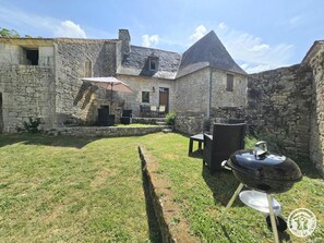 Ein Steinhaus mit einem Rasen, Außenbestuhlung und einem Grill.