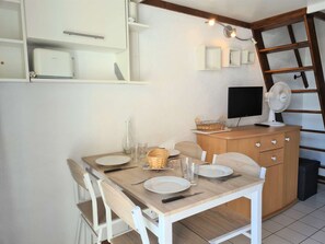 Uma área de refeições com uma mesa de madeira posta para quatro pessoas, uma televisão e um ventilador sobre um armário de madeira.