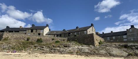A coastal stone building with multiple rooms, a sandy beach in the foreground, and a clear blue sky.