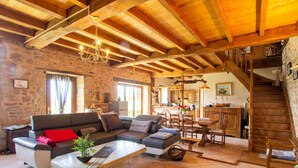 A rustic interior with exposed wooden beams, a stone wall, a sofa, a dining area, and a staircase.