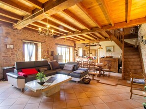 Un interior rústico con vigas de madera a la vista, pared de piedra, un sofá, zona de comedor y una escalera.