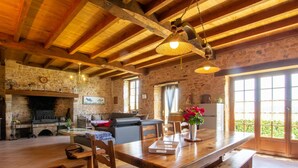 A rustic interior with exposed wooden beams, a stone fireplace, a dining table with benches, and a large window providing natural light.