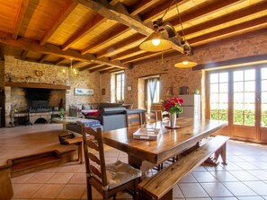 Un interior rústico con vigas de madera a la vista, una chimenea de piedra, una mesa de comedor con bancos y un ventanal que proporciona luz natural.