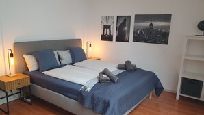 A neatly made bed with a blue quilt, two pillows, and rolled towels. There is a bedside table with a lamp and a power strip. The room has wooden flooring and a white wall with three black-and-white cityscape pictures. - Lichtdurchflutetes Apartment im Herzen der City (Bielefeld)