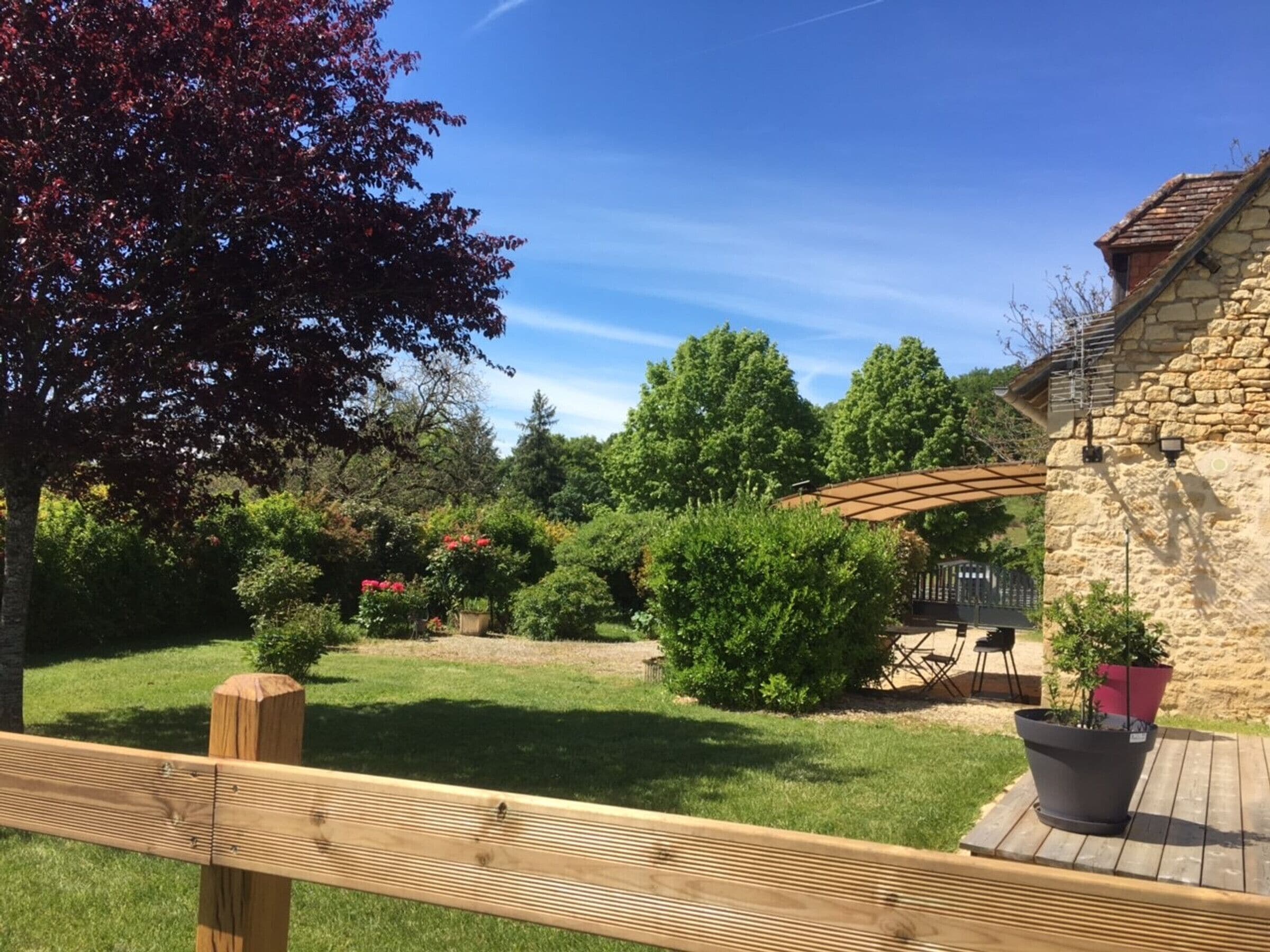 Un terrazzo in legno con un edificio in pietra, un pergolato e un giardino erboso con diverse piante e alberi.