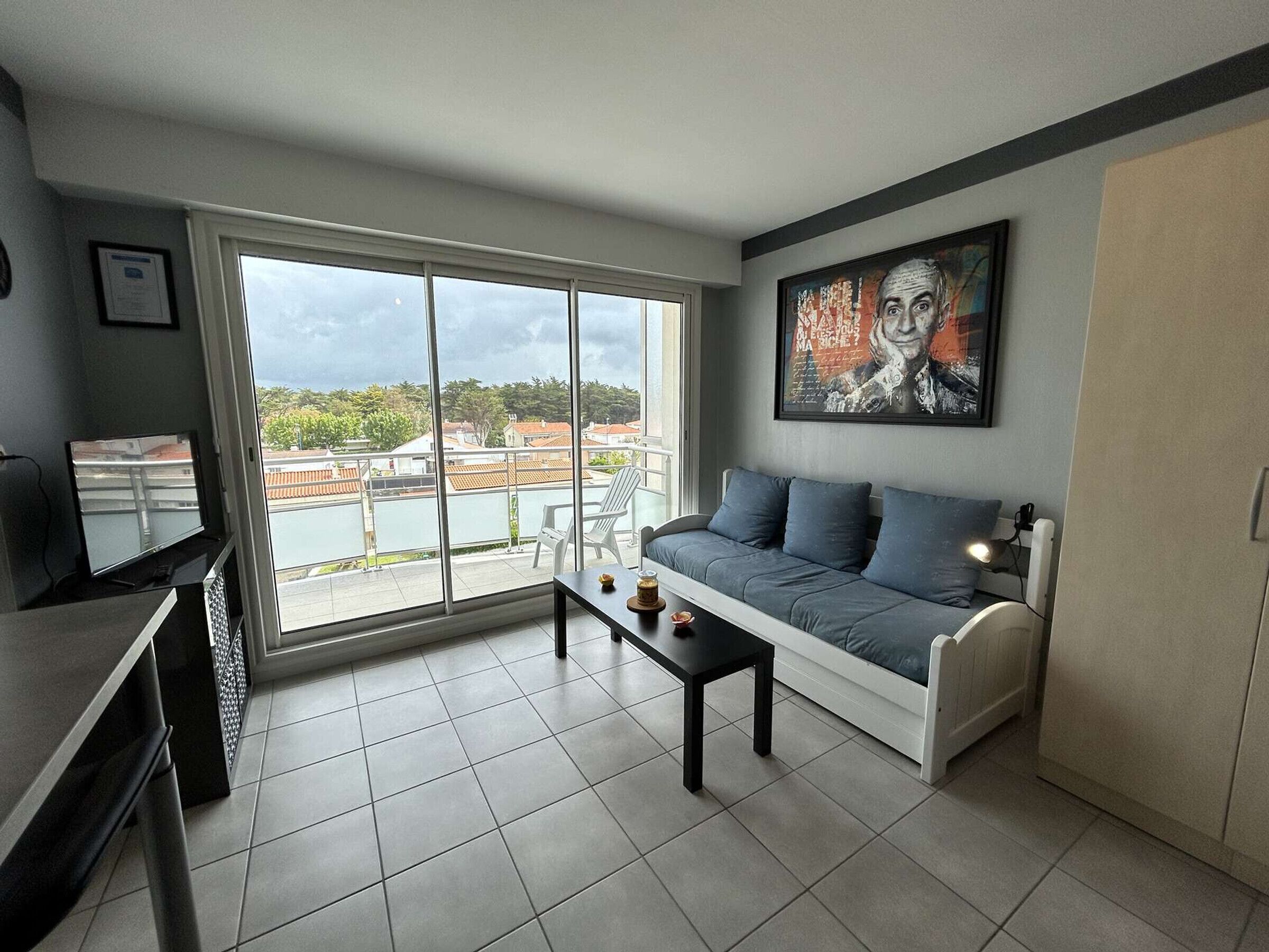 A modern living room with a large sliding glass door leading to a balcony, a sofa with cushions, a small table, and a framed artwork on the wall.