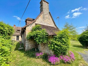 Une charmante maison en pierre, dotée d’un toit en pente, entourée d’une végétation luxuriante et de plantes en fleurs.