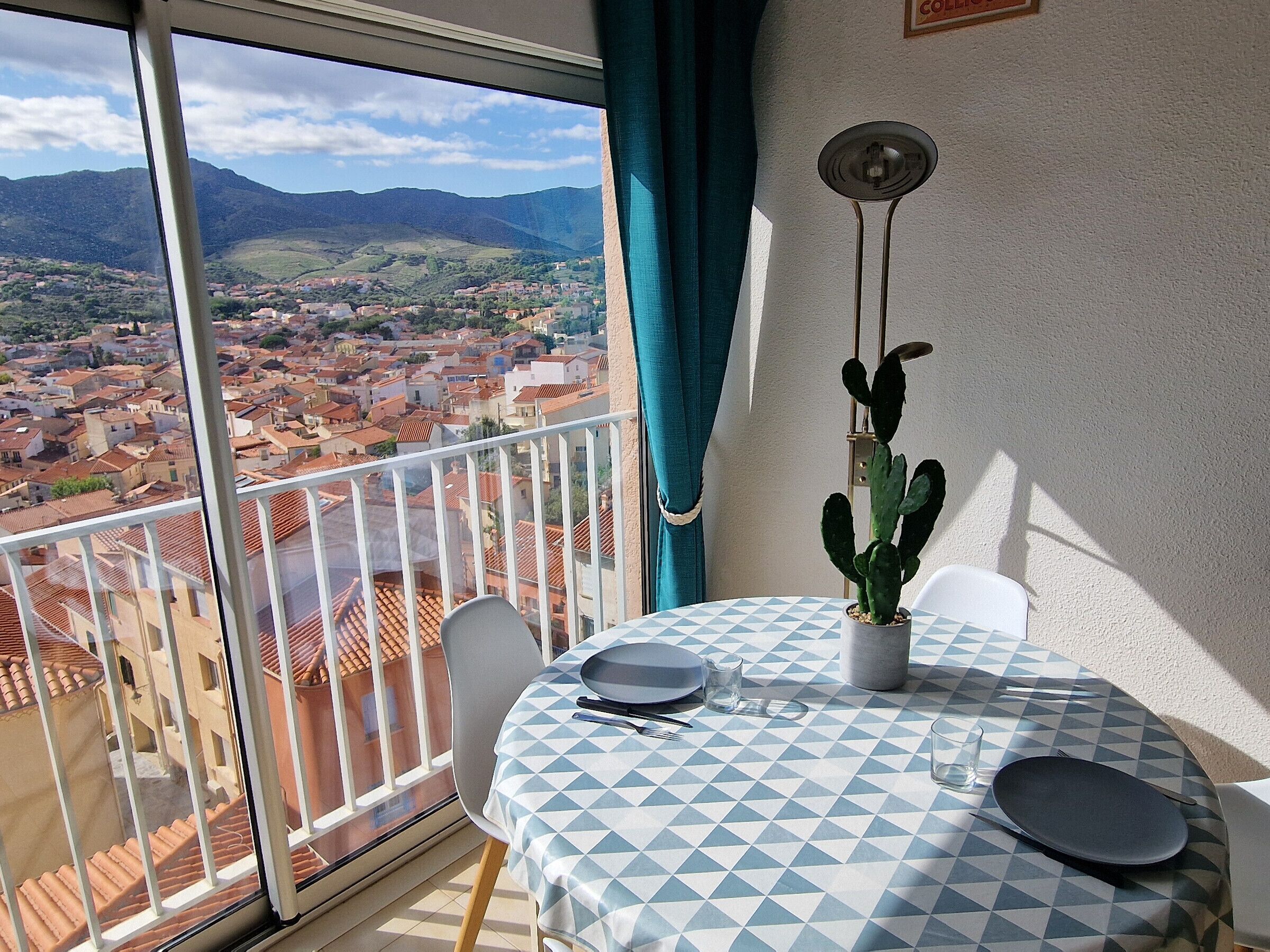 Un balcón con una mesa para dos, un cactus en maceta y vistas de un pueblo con tejados rojos y montañas al fondo.