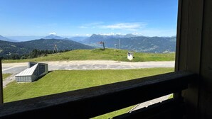 Une vue depuis une fenêtre donnant sur une colline verdoyante, avec un télésiège et des montagnes à l’horizon.