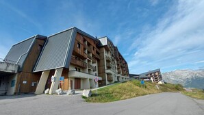 Un bâtiment moderne à plusieurs étages, dont la façade est un mélange de bois et de métal, doté d’un espace de stationnement et surplombant un paysage montagneux.
