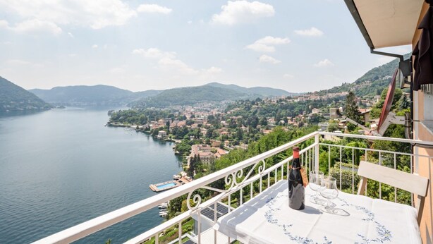 A balcony with a table, a bottle, and glasses, overlooking a lake and a town. - GuestHost - Bright and welcoming studio apartment located in Cernobbio in an elevated position, in a hilly and residential area of the village! The apartment boasts a truly superb lake view: you can admire a unique panorama, which ranges from the swimmin (Cernobbio)