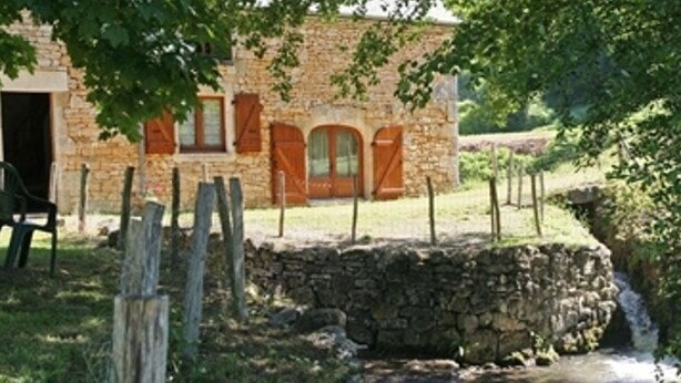 A rustic stone building with a wooden fence, a small stream, and a chair in the foreground.
