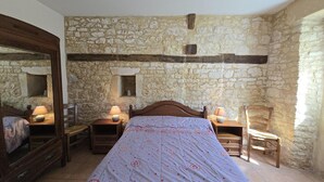 A bedroom with a stone wall, a wooden bed, two bedside tables with lamps, a wooden chair, and a mirror on the left wall.