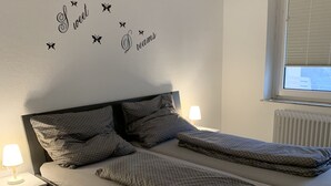 Et soveværelse med to senge, en væg med dekorativ tekst, et vindue med persienner og en sengebordlampe.