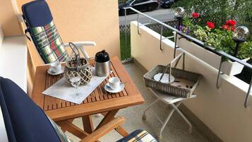 Un balcon avec une table en bois, deux chaises dotées de coussins à carreaux, une bouilloire en métal, un verre et un panier.