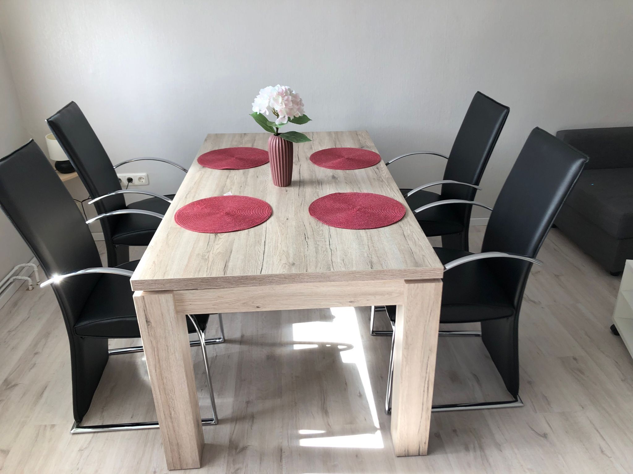 Ein rechteckiger Holztisch mit vier roten Platzsets, umgeben von schwarzen Stühlen mit verchromten Beinen.