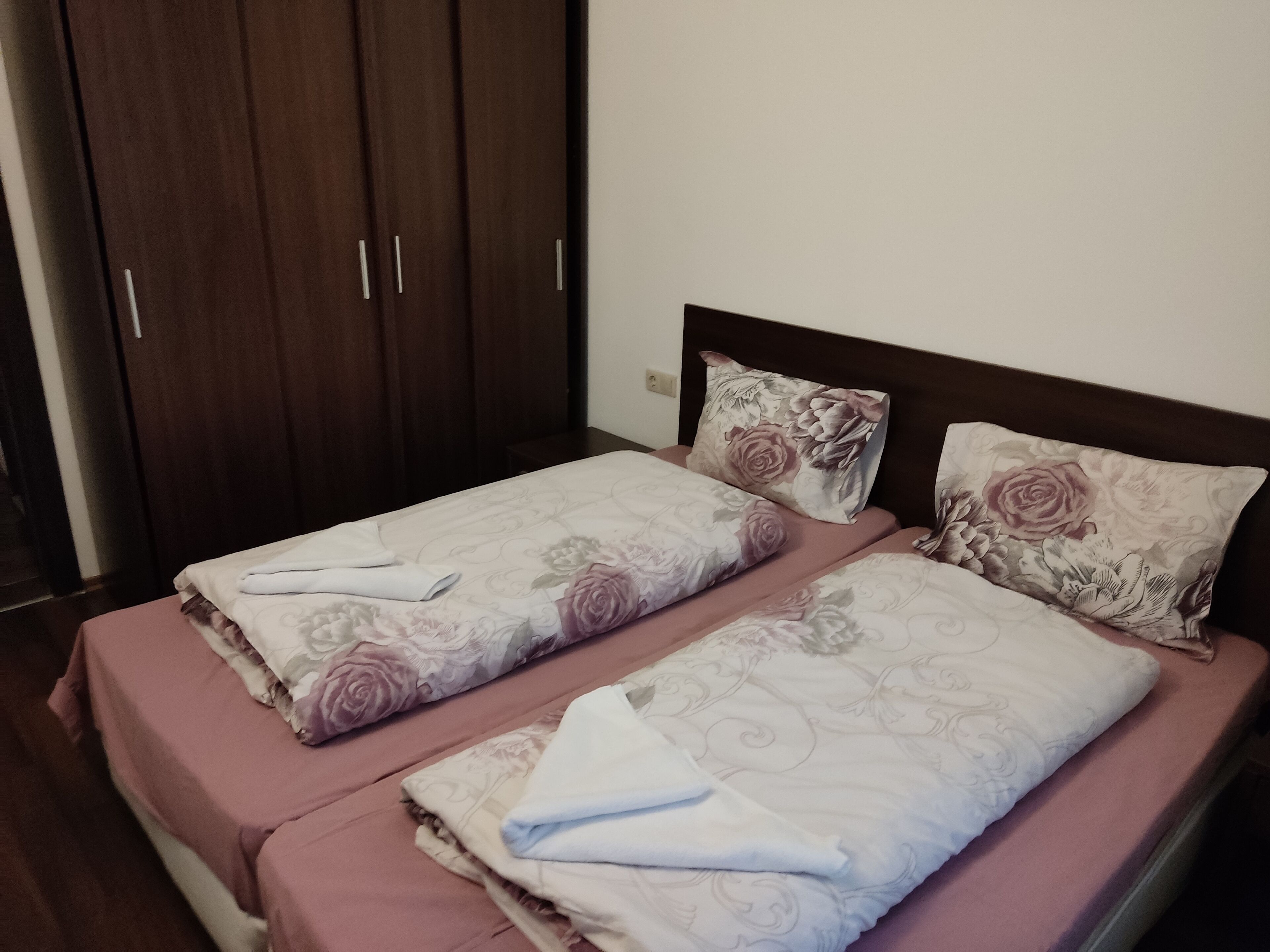 A double bed with floral bedding and two pillows, a wooden wardrobe, and a plain wall.