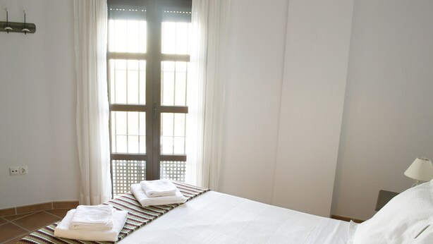 A hotel room with a bed, a window with curtains, a bedside table with a lamp, and a wall socket.