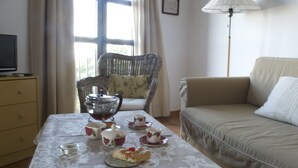 A neatly arranged dining area with a table set for tea, a wicker chair, a sofa, and a television on a dresser.