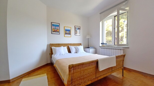 A bedroom with a wooden bed, white bedding, a bedside table with a lamp, and a window with a view of trees. - Holiday apartment Lungomare, only 300m from the beach (Pula)