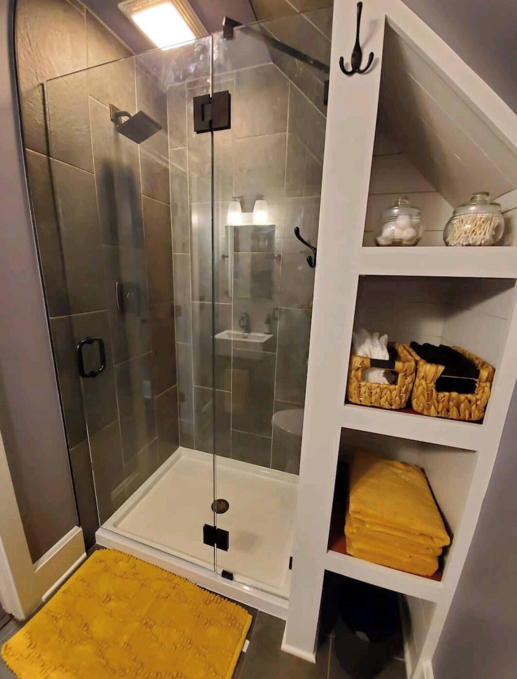 A shower with glass doors, a white shelf with baskets, and a yellow rug.