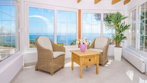 Una habitación iluminada por el sol con muebles de mimbre, una mesa de madera con un florero y amplias ventanas con vista al mar.