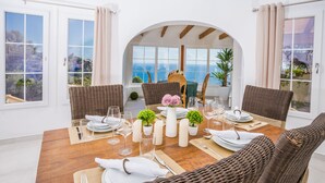 Un comedor con una mesa de madera dispuesta para cuatro personas, rodeado de sillas de mimbre, y vistas al mar a través de amplios ventanales.