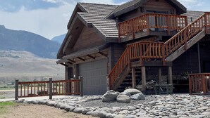 A two-story wooden cabin with a stone foundation, a wooden staircase, and a view of mountains in the background. - Studio 57- luxury apartment in the Wapiti Area! (Cody)
