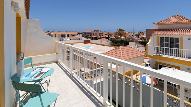 A balcony with turquoise chairs and a table, overlooking a coastal area with buildings and a clear sky. - Modern Apartment with pool in El Castillo - Home For Guest (Antigua - Fuerteventura)