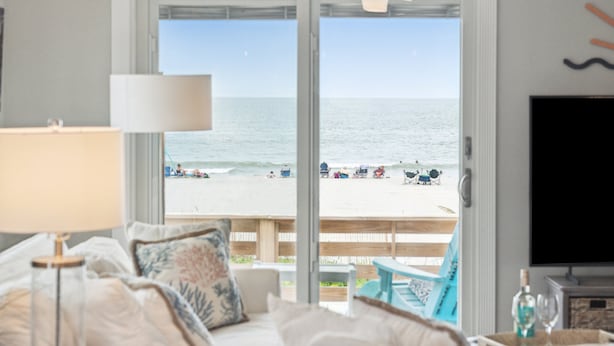 A room with a bed, a TV, a lamp, and a view of the beach. - Oceanfront Luxury Home with Large Deck & Direct Beach Access - No Egrets (Folly Beach)