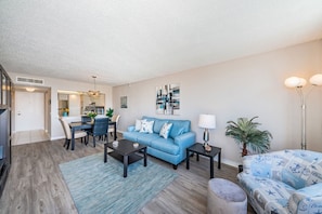 A modern living room with a blue sofa, a dining area with a table and chairs, and a kitchen in the background.