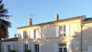 A two-story house with a tiled roof, white shutters, and a small garden with a bench and potted plants. - Maison Individuelle, Preau Vers Piscine Privée Avec Plancha (Saint-Georges-du-Bois)