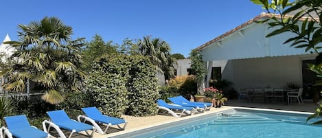 A swimming pool with blue lounge chairs, surrounded by lush greenery and a building with a tiled roof.