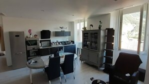 A modern living room with a dining area, kitchen, and a large window providing natural light. - Location Montagne (Veynes)
