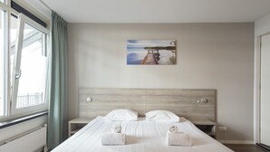 A hotel room with a large bed, two pillows, a nightstand, a painting of a dock, and a window with curtains.
