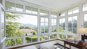 A room with large windows offering a view of a green landscape and distant mountains. There are two cushioned seating areas and a small wooden table with books and candles. - Villa Campo das Medas, in the middle of nature, with private salt pool (Donas Gondomar)