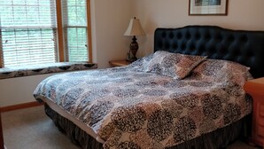 A bedroom with a tufted headboard, a patterned bedspread, a ceiling fan, a bedside lamp, and a window with blinds.