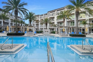 Une piscine entourée de palmiers, avec des chaises longues et des parasols, et un immeuble à plusieurs étages doté de balcons.