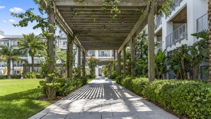 Un paseo techado con suelo de baldosas, rodeado de vegetación y un edificio con balcones.