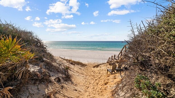 Ein sandiger Strandweg führt zum Meer, mit einer Holzbank und einem Stuhl.